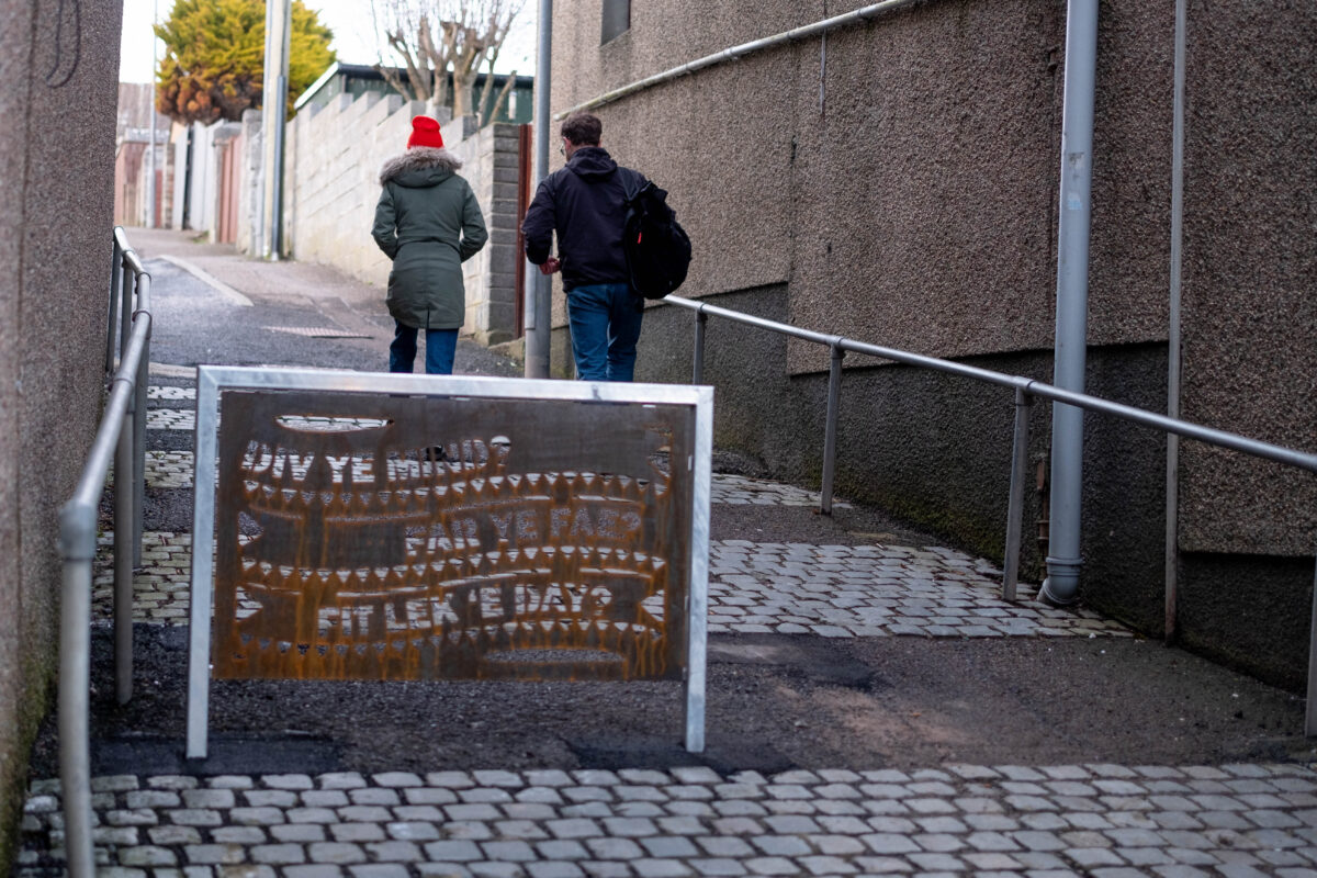 2 adults walk away from camera in background with a poem depicted on a barrier in foreground